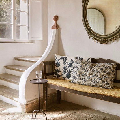 A rustic wooden bench with a yellow cushion sits beside a curved staircase in a sunlit room, decorated with two patterned throw pillows. A large ornate oval mirror hangs on the wall, and a small black side table holds a glass.