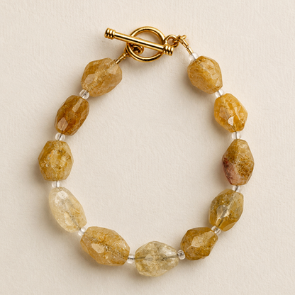 A beaded toggle bracelet featuring faceted amber, yellow, and clear glass beads separated by small silver spacer beads, finished with a gold toggle clasp, shown on a light beige textured background.